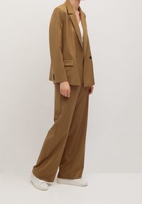 Blazer marron sur mesure et pantalon large assorti, confectionnés en tissu lisse, associés à des baskets blanches. Comprend deux poches avant et des revers.