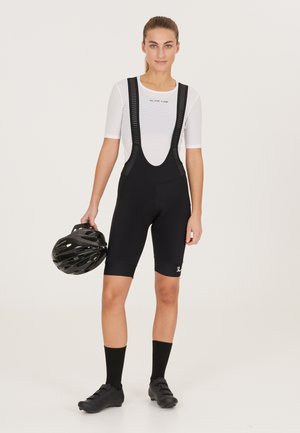 Black cycling bib shorts with mesh straps, short length, and padded seat. Worn with a white moisture-wicking base layer and holding a black helmet.