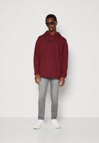 Sudadera granate con un bolsillo frontal y un logo triangular, combinada con jeans gris claro y zapatillas blancas. El modelo lleva gafas de sol.