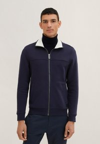 Giacca zip-up blu navy con colletto in pile bianco, texture a costine, due tasche frontali e silhouette aderente. Presenta una cerniera silver.