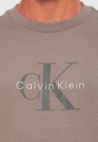 Blizu osobe koja nosi suptilnu mauve majicu s velikim sivim logom "CK" i bijelim logom "Calvin Klein" na prsima.