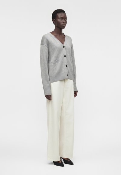 Γυναίκα που φοράει ανοιχτό γκρι ζακέτα με κουμπιά, ψηλόμεσο φαρδύ άσπρο παντελόνι και μαύρες μυτερές γόβες, στέκεται σε λευκό φόντο.