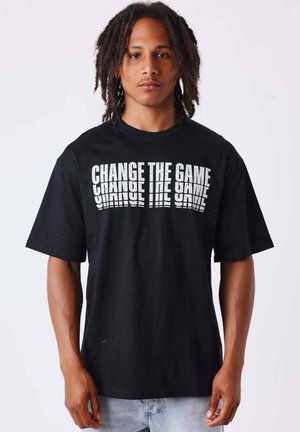 Jonge man met dreadlocks van gemiddelde lengte, die een zwart T-shirt met witte tekst "CHANGE THE GAME" draagt, onderaan gespiegeld.