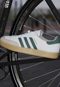 Weißer Sneaker mit grünen Streifen und Akzenten, ausgestattet mit einer strukturierten Gummisohle, einer Wildlederspitze und "VELOSANDA" in Gold geprägt.