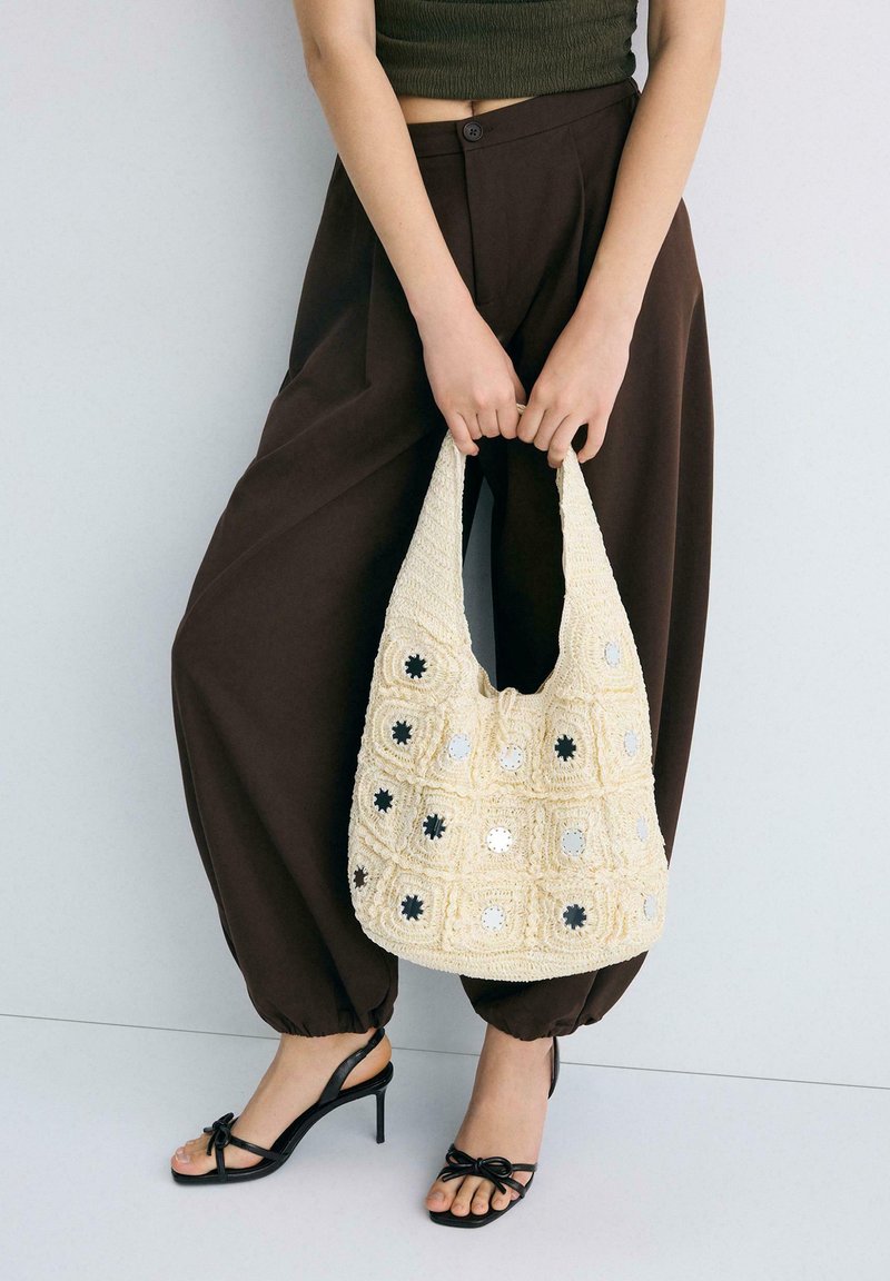 Mains tenant un sac en crochet crème avec des accents de miroir, porté avec un pantalon marron foncé et des sandales à talons ouvertes noires.