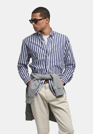 Uomo con occhiali da sole neri, camicia a righe blu e bianca abbottonata, pantaloni beige e felpa grigia legata in vita, guarda a sinistra.