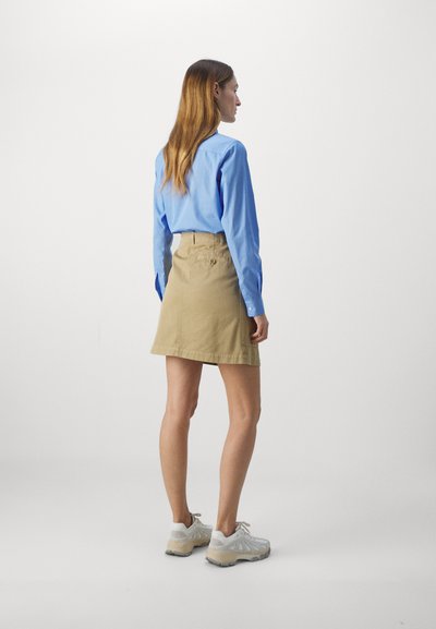 Camicia a maniche lunghe di colore azzurro chiaro abbinata a una gonna A-line beige. Il modello indossa sneakers bianche e grigie. Design semplice e pulito, senza motivi visibili.