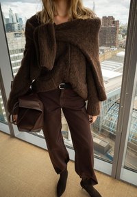 Vrouw draagt een bruine, pluizige trui met bijpassende sjaal, bruine broek, zwarte riem, zwarte laarzen en houdt een donkerbruine leren tas vast, binnen bij een raam.