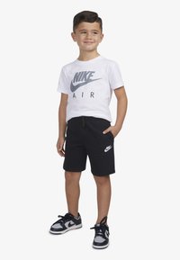 Fehér póló szürke "NIKE AIR" logóval, fekete rövidnadrág oldalzsebekkel és fehér logóval, fekete és szürke edzőcipők, álló pózban.