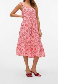 Czerwona, kwiecista midi sukienka z falbaniastym ramiączkiem, guzikiem z przodu i warstwowym dołem. Noszona z dopasowanymi, czerwonymi butami na odkrytej pięcie.