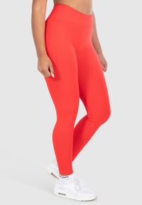 Rode high-waisted leggings van rekbaar materiaal met een gladde textuur en zijspringdetails, gecombineerd met witte sportschoenen.
