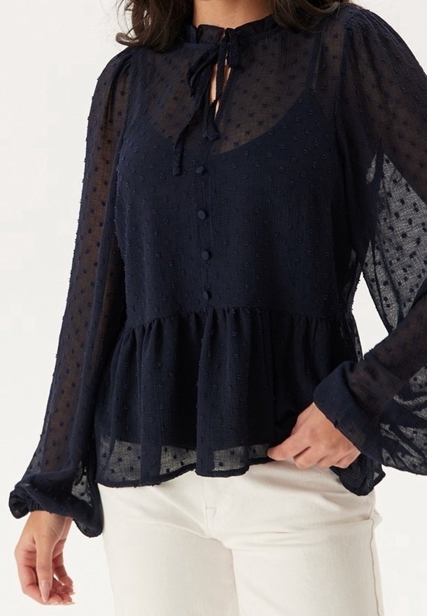 Femme portant un chemisier noir transparent à manches longues avec texture à pois, boutonnage devant, ourlet péplum et col à nouer, associé à un pantalon blanc.