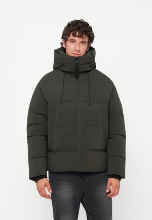 Homme portant une veste matelassée vert foncé avec un col haut et une fermeture éclair devant, debout devant un fond clair uni.