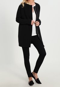 Černý dlouhý kabát s předním zipem, kulatým výstřihem a volným střihem, doplněný o černé kalhoty a ploché boty.