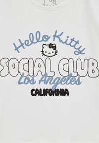 Camiseta blanca con la inscripción "Hello Kitty SOCIAL CLUB Los Ángeles CALIFORNIA" en negro y azul, con un gráfico de Hello Kitty sobre el texto.