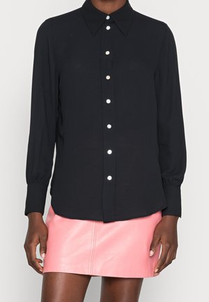 Femme portant un chemisier noir à manches longues avec des boutons en perle et une mini-jupe en cuir rose.