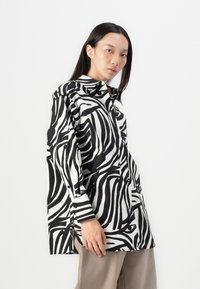 Camicia a righe zebra bianco e nero, con colletto alto, davanti con bottoni, vestibilità ampia e maniche lunghe. Tessuto liscio con finitura lucida.