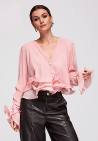 Femme portant un chemisier plissé rose avec des poignets à volants et des boutons dorés, associé à un pantalon en cuir noir et des boucles d'oreilles remarquables.