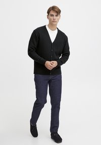 Černý pletený svetr s V-výstřihem, zapínáním na knoflíky a žebrovanými manžetami, zkombinovaný s tmavými džíny a černou obuví, stojící na bílém pozadí.