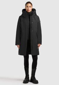 Svart parka med huva, sidofickor och dragkedja; lagd över en matchande svart puffjacka. Slät textur och elegant design.