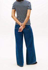 Personne debout, dos à la caméra, vêtue d’une chemise à manches courtes rayée bleu marine et blanc et d’un jean bleu à jambes larges.