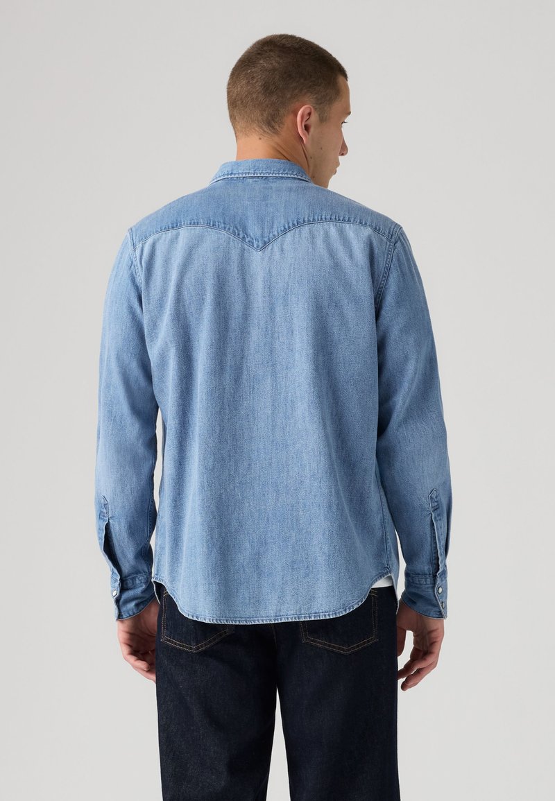 Homme de dos portant une chemise en denim bleu clair avec des poignets boutonnés et un jean bleu foncé sur un fond gris uni.