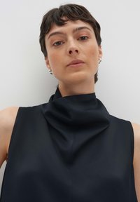 Top nero senza maniche con colletto alto drappeggiato, realizzato in tessuto morbido. La modella ha i capelli corti e indossa piccoli orecchini a cerchio.