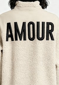 Veste en polaire beige avec une surface texturée et douce, ornée d'un "AMOUR" noir brodé dans le dos. Le col est plat et minimaliste.