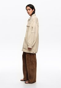 Young woman with shoulder-length dark hair wears oversized beige jacket, wide brown pants, and black sandals, standing in profile.