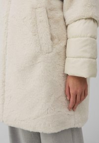 Crème faux fur jas met een pluche textuur, voorzien van een puffy, gladde ivoorkleurige mouw en een zijzak. De onderrand is afgerond.