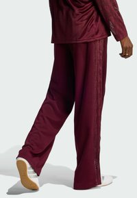 Pantalon de sport bordeaux à coupe décontractée, avec des bandes latérales et une texture douce. Associé à des chaussures de sport blanches.