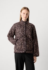 Woman wearing a black floral quilted jacket with round silver buttons and large curved front pockets, paired with black pants.