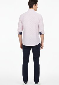 Hombre que lleva una camisa de manga larga de color rosa claro con estampado, mangas remangadas, pantalones azul marino oscuro y zapatillas blancas, de pie mirando hacia atrás sobre un fondo blanco.