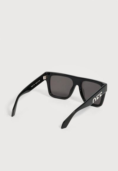 Lunettes de soleil rectangulaires noires avec des verres foncés et un texte blanc sur la branche droite visible sur fond blanc.