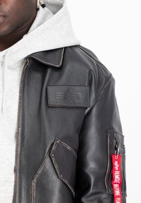 Svart läderjacka med en framträdande patch med logotyp, kontrasterande sömmar och en röd nyckelringsetikett med text. Under jackan finns en grå hoodie.