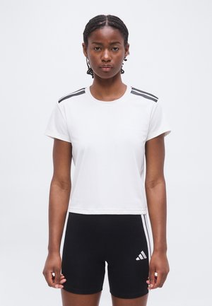 Mujer con cabello trenzado con una camiseta blanca corta con rayas negras en los hombros y pantalones cortos negros con rayas blancas y logo.