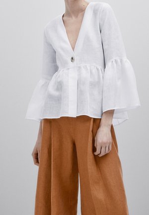 Femme portant un chemisier blanc à col en V avec des manches évasées et un seul bouton, assorti à un pantalon large marron taille haute.