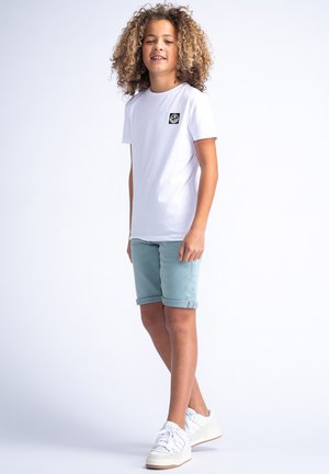 Witte T-shirt met korte mouwen en zwart logo op de borst, lichtblauwe shorts met opgerolde zoom en witte sneakers. Glad materiaal, casual pasvorm.