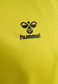 Gelbes Sportshirt mit einem schwarzen Logo, das ein stilisiertes Bienenmotiv und den Text "hummel" zeigt. Glatte Stoffoberfläche mit strukturiertem Finish.