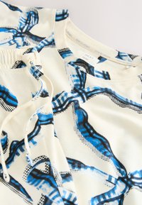 Haut en coton avec un fond crème orné de motifs abstraits bleus et noirs. L'encolure est ronde avec un cordon de serrage à l'ourlet.