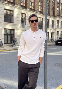 Ung mand iført solbriller, hvid henley-skjorte og mørke bukser læner sig op ad en gadepæl på en byfortov med cykler parkeret i nærheden.