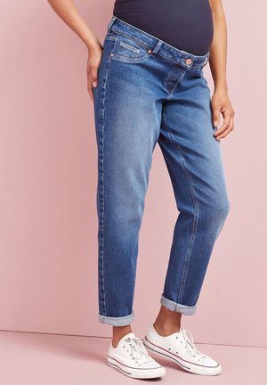 Pregnant person wearing loose-fitting blue maternity jeans with rolled cuffs and white sneakers against a pink background.