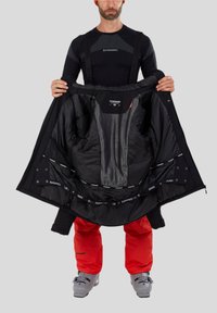 Černá izolovaná bunda s podrobnou podšívkou; červené lyžařské kalhoty; osoba držící bundu otevřenou, aby ukázala její vnitřní prvky a design.