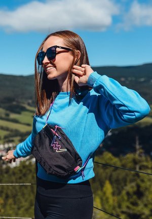 Black crossbody bag with colorful geometric accents, worn over a blue sweatshirt. Model stands outdoors with a green landscape in the background.