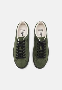 Gröna mockaskor med svarta snören, rund tå och kontrasterande vit kant. Har en logotyp tryckt på innersulan. Platt gummisula.