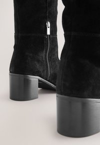 Schwarze Wildleder-Stiefeletten mit Blockabsätzen und seitlichen Reißverschlüssen vor hellem Hintergrund, von hinten und von der Seite betrachtet.