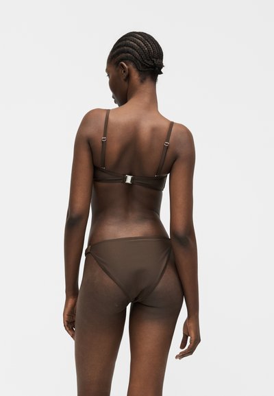 Γυναίκα με πλεγμένα μαλλιά που φοράει καφέ μπικίνι, όρθια με την πλάτη στην κάμερα σε ένα απλό λευκό φόντο.