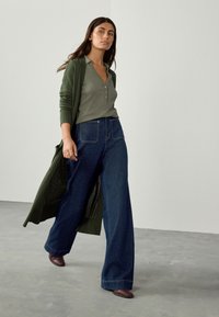 Cardigan verde, maglietta henley grigia, jeans a gamba larga blu scuro e scarpe bordeaux. Tessuto liscio, vestibilità rilassata, silhouette lunga.