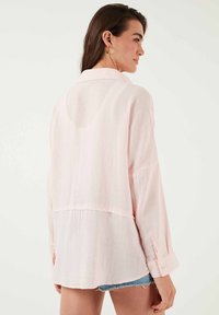 Light pink, loose-fitting shirt in textured fabric with a collar, long sleeves, and a hemline that drops at the back.