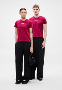 Dva modelové mají na sobě shodná vínově červená trička s krátkým rukávem a nápisem "TOMMY", kombinovaná s širokými černými kalhotami. Jeden drží malou černou kabelku.